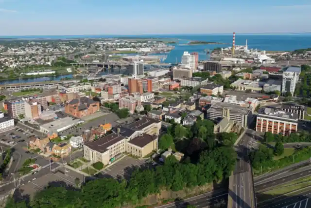 Aerial view of downtown Bridgeport with a highway in the foreground, the harbor and a red-and-white smokestack behind, and Long Island Sound on the horizon
